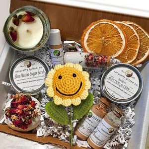 May include: A gift box filled with self-care items. Includes a candle with rose petals, shea butter sugar scrub, shea butter lotion, dried orange slices, and a crocheted sunflower. The box also contains small bottles and a bath bomb.