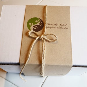 May include: A white cardboard box with a brown paper band tied with twine. The band features a logo with a green and brown yin-yang design and the text "Naturally Gifted". The twine is tied in a bow.