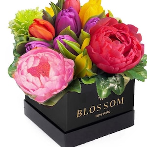 May include: A square black box with the gold text BLOSSOM NEW YORK printed on the front, containing a vibrant floral arrangement of pink and red peonies, purple, yellow, and orange tulips, and light green carnations with green leaves.