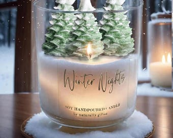 May include: A lit soy hand-poured candle in a clear glass jar, featuring three green Christmas tree-shaped candles. The candle is labeled "Winter Nights" and sits on a wooden coaster with a snowy rim. The background shows a snowy forest.