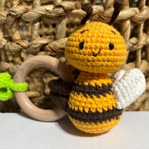 May include: A handmade crochet bee baby rattle. The bee features a yellow body with brown stripes, white wings, and a smiling face. It has a wooden ring and a green crochet accent. The background includes a woven basket.