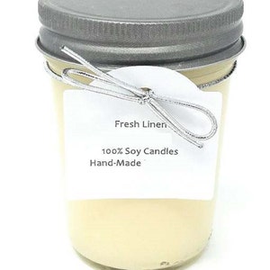May include: A clear glass jar candle with a silver lid and a silver wire bow. The label reads "Fresh Linen", "100% Soy Candles", and "Hand-Made". The candle has a light yellow color.