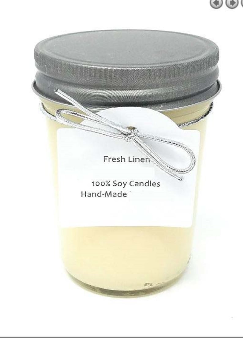 May include: A clear glass jar candle with a silver lid and a silver wire bow. The label reads "Fresh Linen", "100% Soy Candles", and "Hand-Made". The candle has a light yellow color.