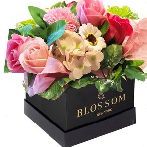 May include: A black square box with a gold logo that reads "BLOSSOM NEW YORK". The box is filled with a bouquet of pink, white, and red soap flowers.