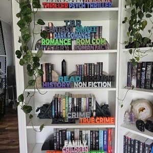 Pode incluir: Uma estante branca com v&aacute;rias prateleiras cheias de livros. Os livros est&atilde;o organizados por g&ecirc;nero, com blocos de madeira coloridos que soletram os nomes dos g&ecirc;neros. Os g&ecirc;neros incluem Young Adult, Horror, Thriller, Fantasy, Smut, Dark Romance, Romance, Rom Com, Mystery, Romantacy, TBR, Read, Biography, History, Non Fiction, Sci Fi, Crime, True Crime, Family, Children, Fiction, and Adventure.