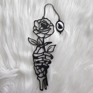 May include: Black metal wall art featuring a rose held by a skeleton hand. A small oval frame with a raven silhouette hangs from a chain. The artwork is set against a white, fluffy background.
