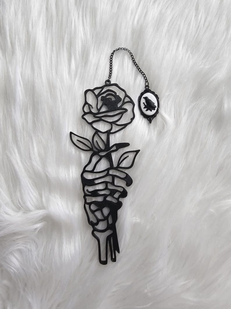 May include: Black metal wall art featuring a rose held by a skeleton hand. A small oval frame with a raven silhouette hangs from a chain. The artwork is set against a white, fluffy background.