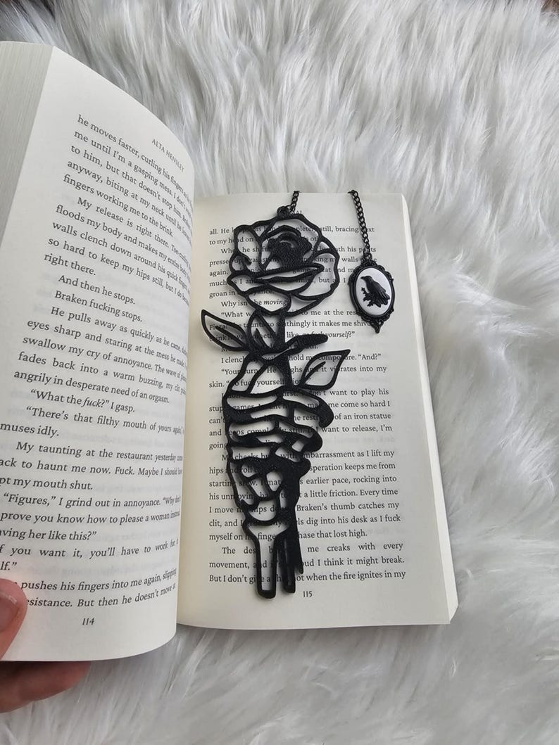 May include: Black metal rose and raven bookmarks resting on the pages of a book. The rose bookmark features a detailed floral design, while the raven bookmark is in an oval frame. The book is open to a page with printed text.