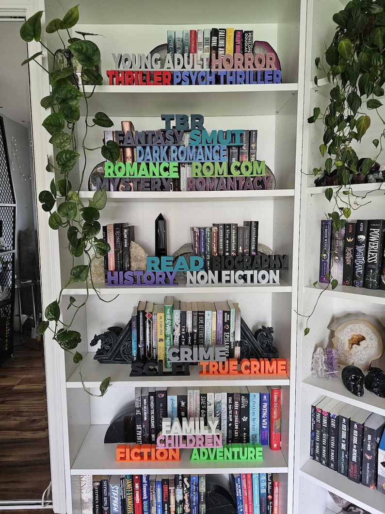 Pode incluir: Uma estante branca com v&aacute;rias prateleiras cheias de livros. Os livros est&atilde;o organizados por g&ecirc;nero, com blocos de madeira coloridos que soletram os nomes dos g&ecirc;neros. Os g&ecirc;neros incluem Young Adult, Horror, Thriller, Fantasy, Smut, Dark Romance, Romance, Rom Com, Mystery, Romantacy, TBR, Read, Biography, History, Non Fiction, Sci Fi, Crime, True Crime, Family, Children, Fiction, and Adventure.