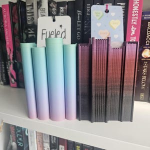 May include: A bookshelf with a variety of books, including "Twist Me", "Never Let Go", "Honestly I'm Too", and "Fueled". There are also several bookends, including a set of four ombre bookends in shades of blue and pink, and a set of bookends in shades of red and black.