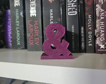 May include: A sparkly, magenta-colored ampersand symbol, standing on a shelf with a row of books behind it. The books have titles like "Soul for Revenge" and "Bloodshed".