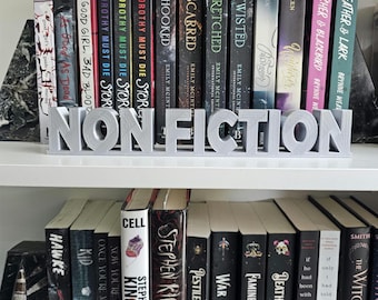 May include: A bookshelf with a silver 3D word "NONFICTION" bookend. The bookshelf is filled with books, some with titles visible, including "Good Girl, Bad Blood", "Twisted", "Scarred", "Hooked", "Cell", "Stephen King", "Pestillence", "War", "Famine", "Death", "The Witch Haven", and "Once You're Mine".