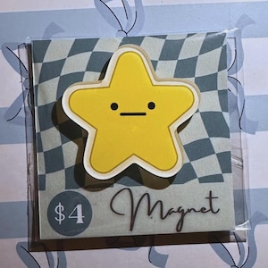 Peut inclure: Un aimant en forme d'étoile jaune avec un contour blanc et un visage simple. L'aimant est sur une carte avec un motif à carreaux et le mot "Magnet". Le prix est de 4 $.