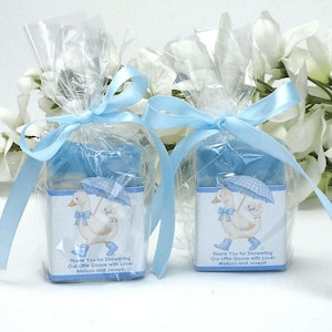 Two blue silly goose baby shower soap favors wrapped in clear gift packaging with soft blue ribbons, featuring handmade blue soap bars and matching goose design labels, styled with white floral accents in the background