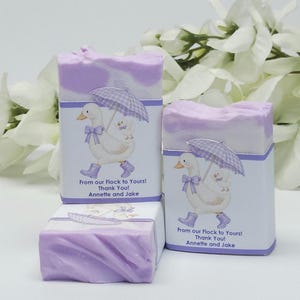 Lavender silly goose baby shower soap favors displayed together, showing two wrapped handmade lavender soap bars with goose design labels and one unwrapped soap, styled with white floral accents in the background