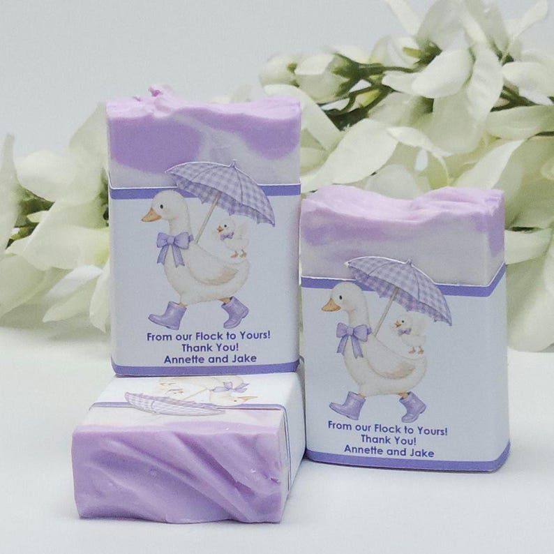 Lavender silly goose baby shower soap favors displayed together, showing two wrapped handmade lavender soap bars with goose design labels and one unwrapped soap, styled with white floral accents in the background