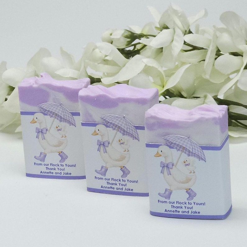 Three lavender silly goose baby shower soap favors displayed unwrapped, showing handmade lavender and cream soap bars with matching goose design labels, styled with white floral accents in the background