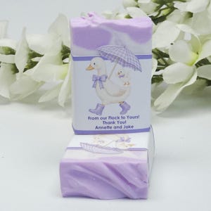Lavender silly goose baby shower soap favor displayed with one wrapped handmade lavender soap bar featuring a goose design label and one unwrapped soap, styled with white floral accents in the background
