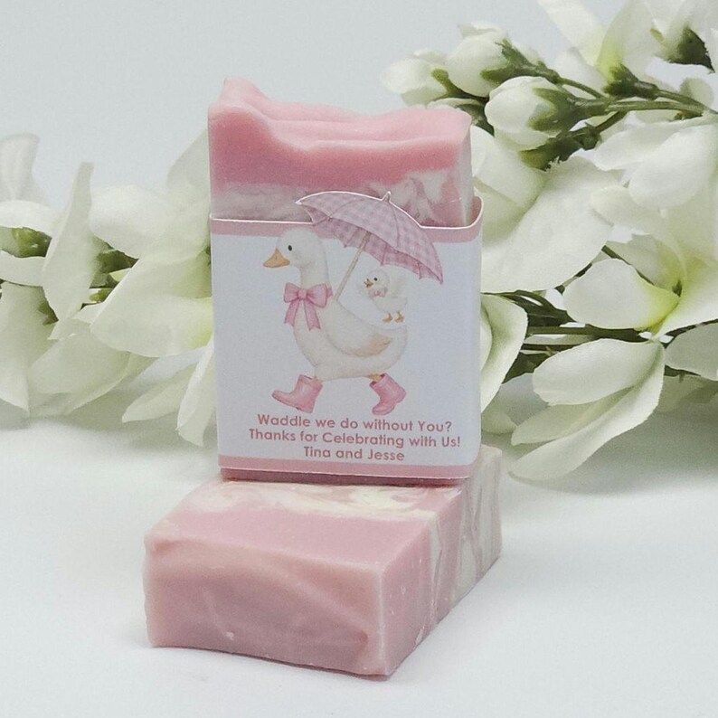 Pink silly goose baby shower soap favor displayed with one wrapped handmade pink soap bar featuring a goose design label and one unwrapped soap, styled with white floral accents in the background