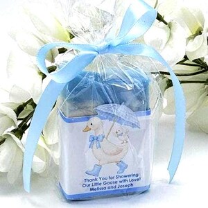 May include: A baby shower favor wrapped in clear cellophane and tied with a light blue ribbon. The favor features a white box with a duck and baby duck illustration, holding an umbrella. Text reads: "Thank You for Showering Our Little Goose with Love! Melissa and Joseph."