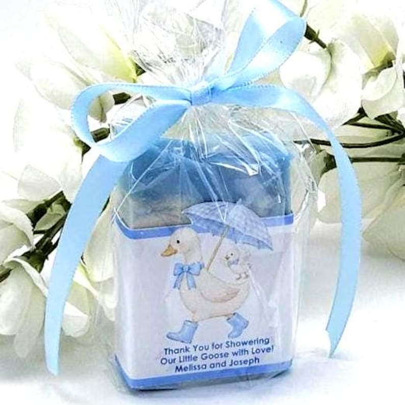 May include: A baby shower favor wrapped in clear cellophane and tied with a light blue ribbon. The favor features a white box with a duck and baby duck illustration, holding an umbrella. Text reads: "Thank You for Showering Our Little Goose with Love! Melissa and Joseph."
