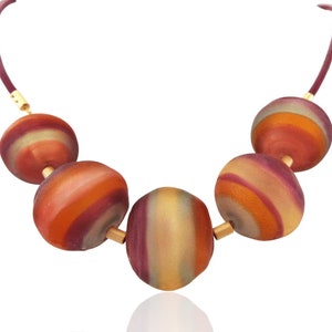May include: A necklace featuring five large, round beads in shades of orange, red, and yellow with a striped pattern. The beads are strung on a dark red cord with gold-colored metal accents. The necklace is a statement piece.