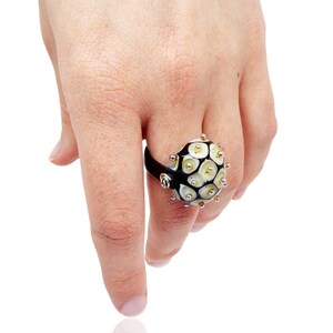 May include: A black ring with a white glass sphere featuring a textured design of small circles. The sphere is accented with silver and gold dots.