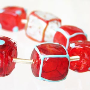 May include: A close-up of a red and blue glass bead necklace. The beads are cube-shaped and have a white outline. The necklace is strung on a silver wire.