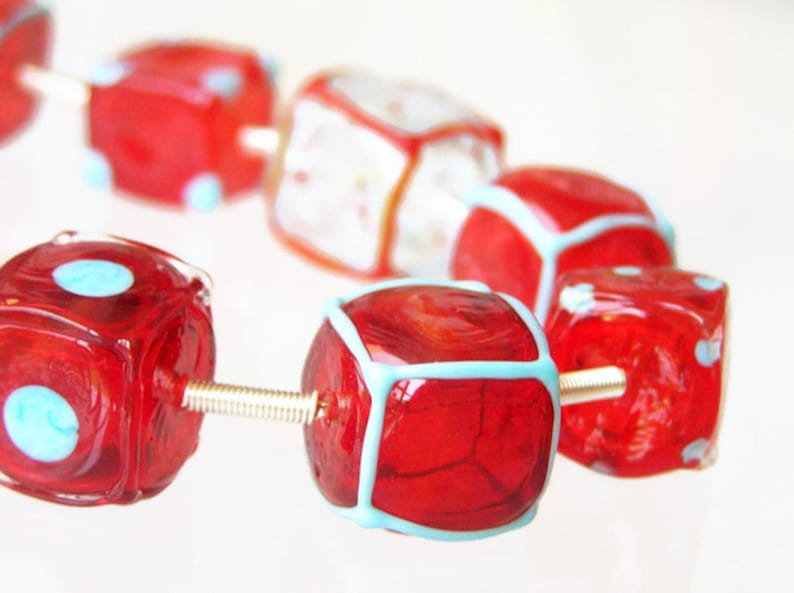 May include: A close-up of a red and blue glass bead necklace. The beads are cube-shaped and have a white outline. The necklace is strung on a silver wire.