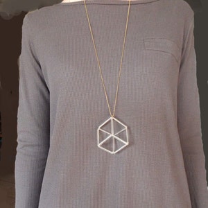 May include: A long gold chain necklace with a geometric pendant. The pendant is a silver-coloured hexagon with an internal starburst design. The necklace hangs over a grey long-sleeved shirt.