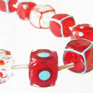 May include: A strand of red and white glass beads in the shape of dice. Some of the beads have a blue outline and white polka dots.