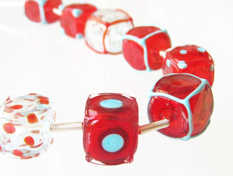 May include: A strand of red and white glass beads in the shape of dice. Some of the beads have a blue outline and white polka dots.