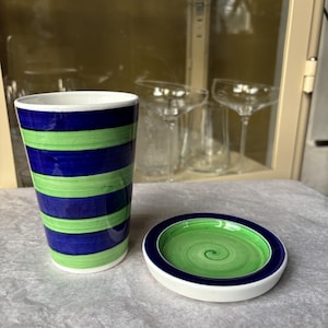 Könnte beinhalten: Ein Keramikbecher und ein passendes Untertassen-Set. Der Becher hat horizontale Bänder in Blau und Grün. Die Untertasse hat ein grünes Spiraldesign innerhalb eines blauen Randes. Beide Teile haben einen weißen Sockel.