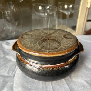 以下が含まれることがあります： 丸い陶器製の蓋付き容器で、外側は濃い茶色、蓋は明るい茶色です。蓋には円形の模様と赤褐色の縁があります。容器には2つの小さなハンドルがあり、白い表面に置かれています。