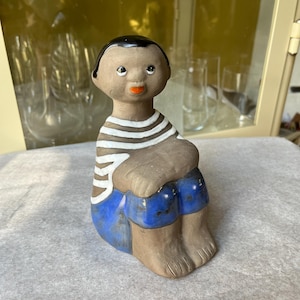 May include: A ceramic figurine of a seated figure with dark hair and skin. The figure wears a striped shirt, blue shorts, and has their arms wrapped around their knees. The figurine has a simple, folk art style.