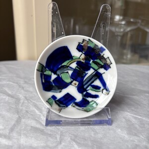 May include: A small, round, white ceramic dish with an abstract design in shades of blue, green, and brown. The dish is displayed on a clear plastic stand, showcasing its decorative pattern. The dish is likely for serving small portions.