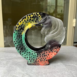 May include: A circular vase with a horse head design. The vase is painted with a gradient of colours, including green, yellow, and red, with black speckles. The horse head is a frosted, translucent white.
