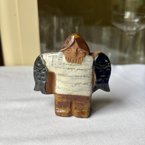 Könnte beinhalten: Keramikfigur einer Person, die zwei Fische hält. Die Figur hat einen braunen Kopf und Beine, ein weißes Hemd mit horizontalen Linien und zwei dunkelblaue Fische. Die Figur steht auf einer weißen Oberfläche.