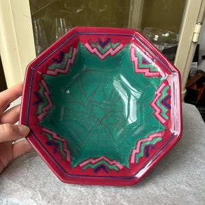 Op de afbeelding: Een achthoekige keramische kom met een rode rand en een groene binnenkant. De binnenkant heeft een rood, webachtig patroon. De rand is versierd met roze, blauwe en paarse geometrische vormen. Een decoratief stuk om te serveren of tentoon te stellen.
