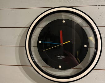 May include: A round wall clock with a black face, white and black frame, and clear glass. The clock has a red second hand, a yellow minute hand, and a blue hour hand. The clock face has the text "VERICHRON QUARTZ".