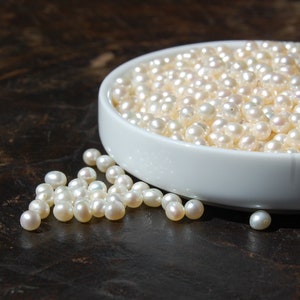 May include: A white ceramic bowl filled with white pearls. A scattering of pearls is on a dark brown surface.