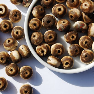 Könnte beinhalten: Eine Sammlung brauner und weißer Knochenperlen mit einem Loch in der Mitte jeder Perle. Die Perlen befinden sich in einer weißen Schale und sind auf einer weißen Oberfläche verstreut.