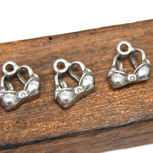 May include: Three silver charm pendants shaped like brassieres. The charms are on a wooden surface.