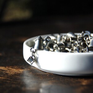 Puede incluir: Un brazalete de plata con un dije de cafetera colgando. El brazalete está en un tazón de cerámica blanco.