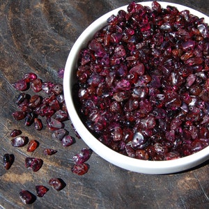 May include: A white bowl filled with small, dark red, irregularly shaped gemstones. Some of the gemstones are scattered on a dark brown wooden surface.