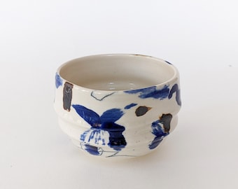 May include: A ceramic bowl with a white base and blue and brown abstract floral designs. The bowl has a textured, ridged exterior and a smooth interior. The rim is slightly flared, and the bowl is suitable for serving or display.
