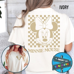 May include: A white t-shirt with a checkered pattern and a gold Minnie Mouse graphic on the back. The text "Minnie Mouse" is printed below the graphic. The shirt is available in ivory.