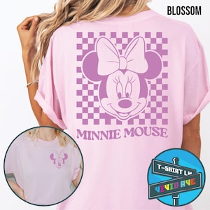 May include: Pink t-shirt with a checkered pattern and a purple Minnie Mouse graphic. The text "Minnie Mouse" is printed below the graphic. The t-shirt is available in sizes S-XL.