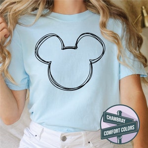 Puede incluir: Camiseta azul claro con un contorno negro de la cabeza de Mickey Mouse. La camiseta tiene mangas cortas y cuello redondo. Los textos "CHAMBRAY" y "COMFORT COLORS" est&aacute;n en un letrero en la esquina inferior derecha.