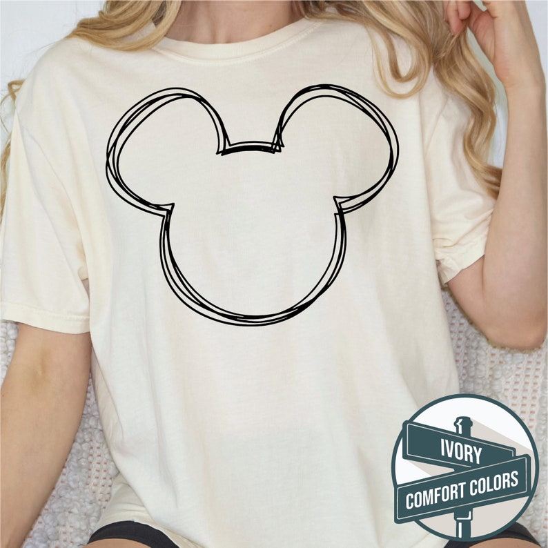 Puede incluir: Camiseta de color marfil con un contorno negro de la cabeza de Mickey Mouse. El dise&ntilde;o es de estilo simple y esbozado. La camiseta forma parte de la colecci&oacute;n Comfort Colors.